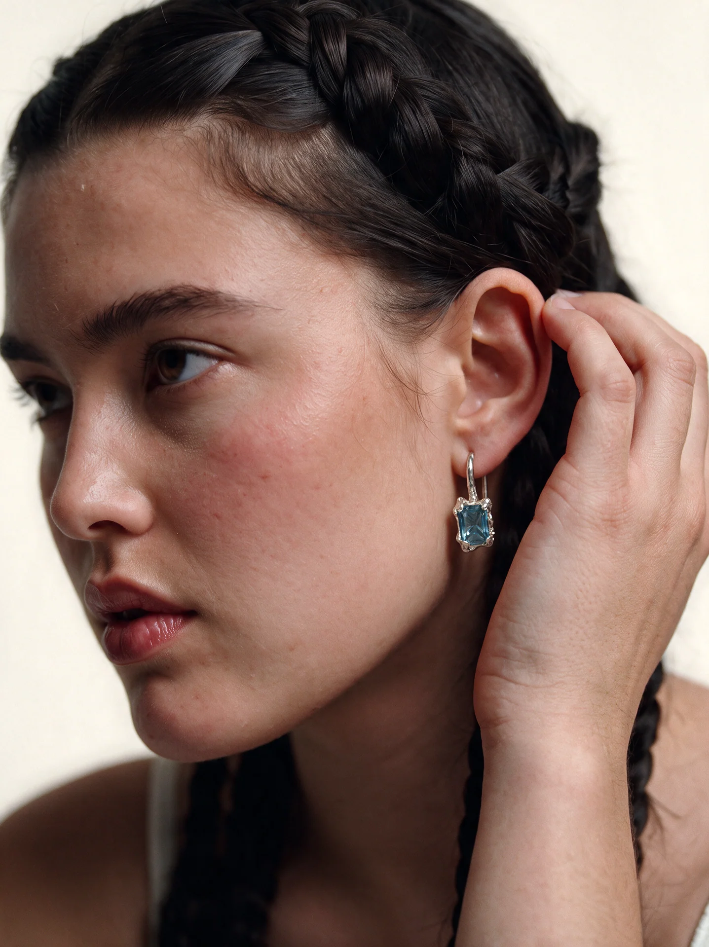 Close portrait of a model with braided hair wearing a silver-toned earring with a blue centerpiece