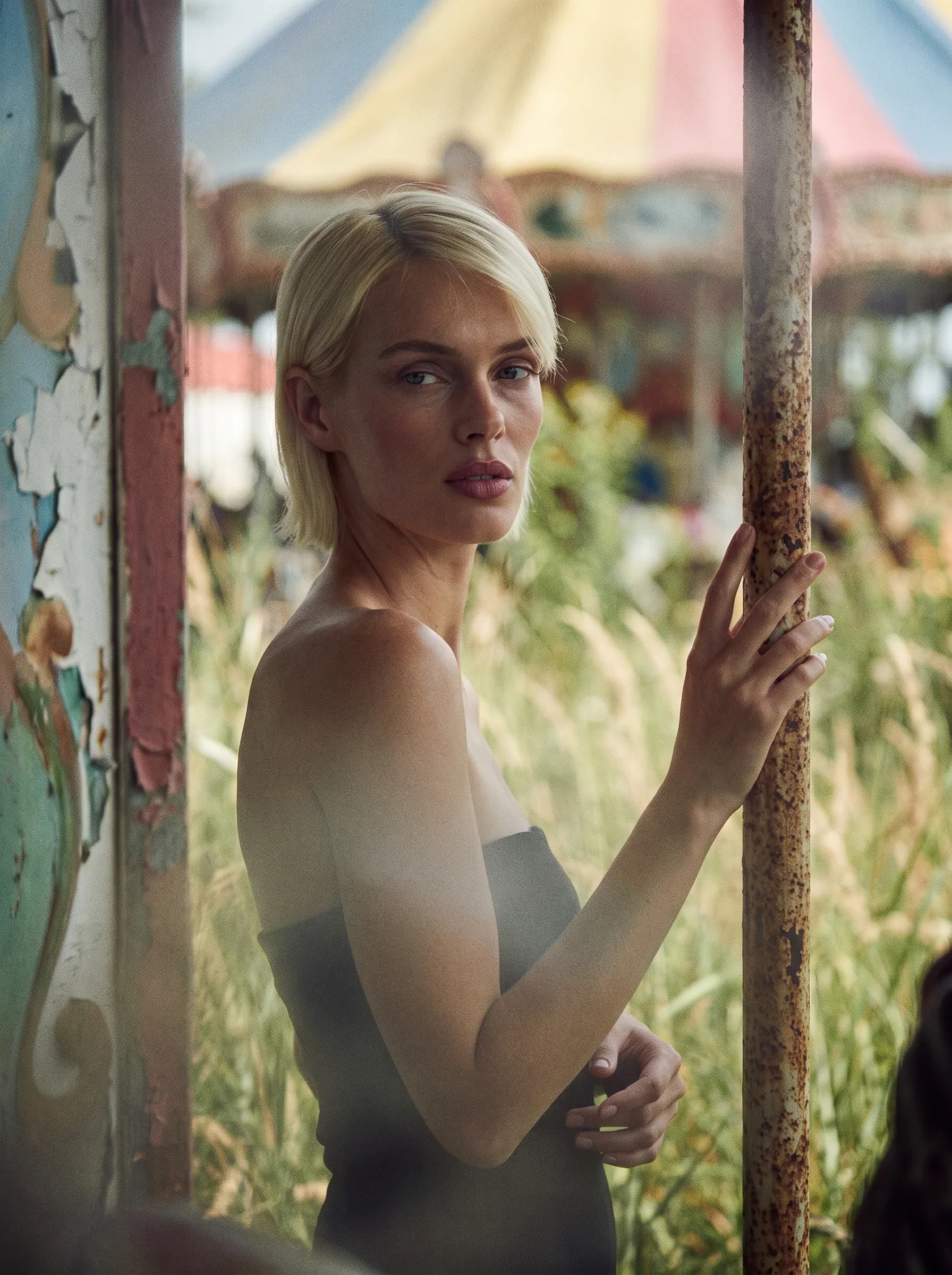 Blonde model in a black dress standing beside a rusted post at a weathered fairground