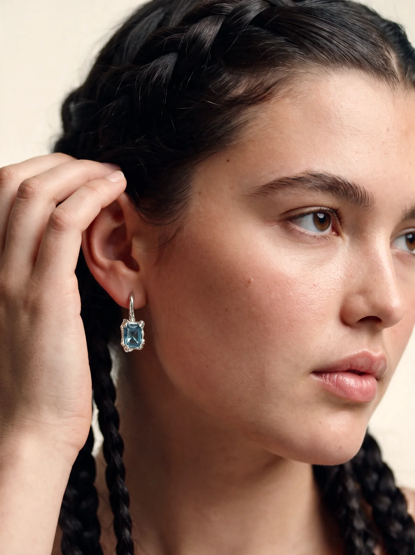 Model with braided hair touching her ear while wearing a silver-toned blue earring